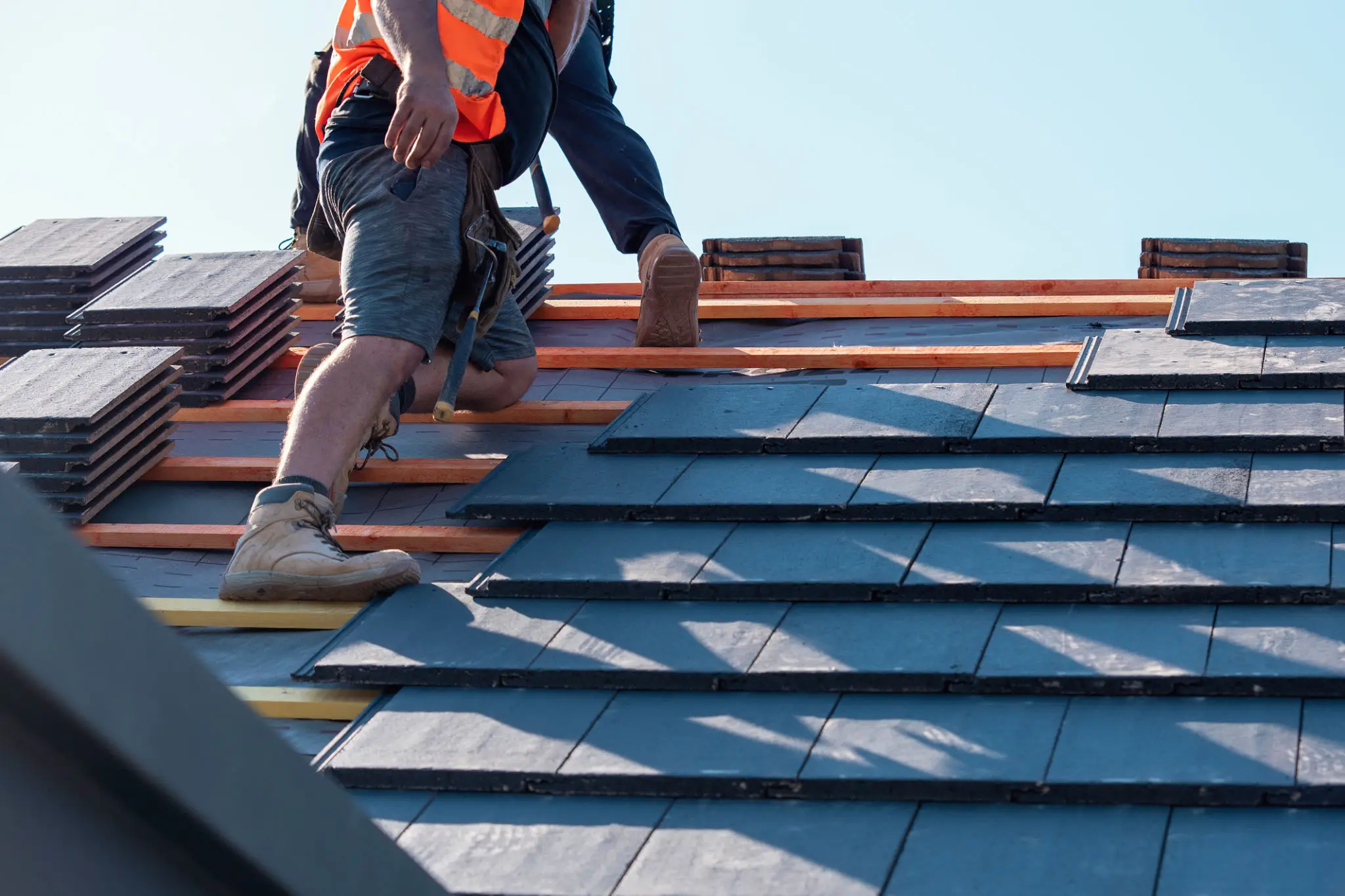 Workers installing slate tiles on a roof under clear sky.
